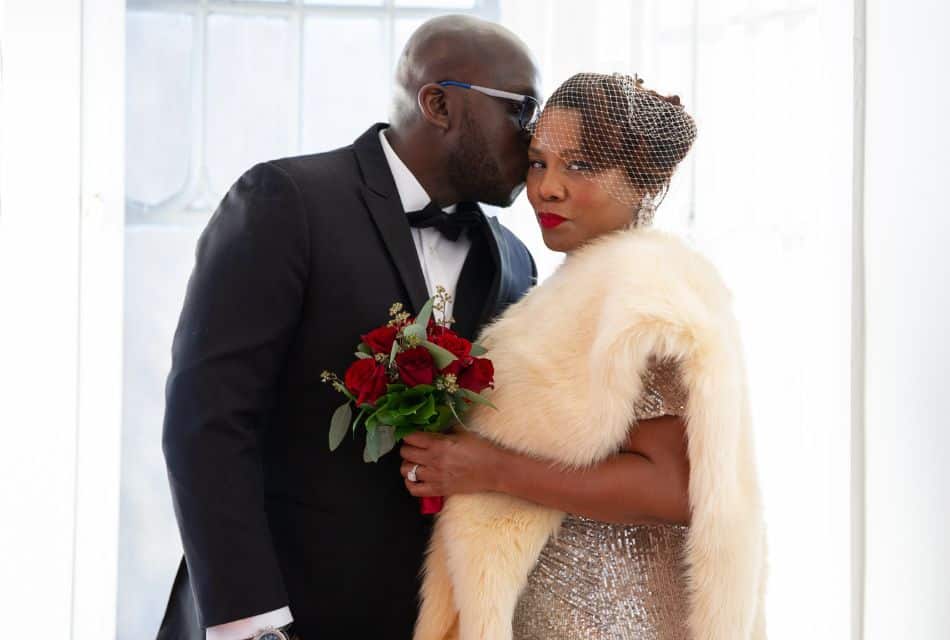 A woman in a sparkling gold dress, fur shawl and netted hat standing beside a man in a black tuxedo and sunglasses